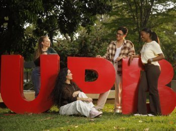 Cuatro jóvenes posan sonrientes al aire libre junto a las grandes letras rojas "UPB" sobre el césped, con árboles al fondo.