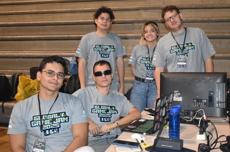 Grupo de jóvenes en un puesto de videojuegos, con camisetas grises y lanyards, trabajando frente a computadoras y monitor en un evento.