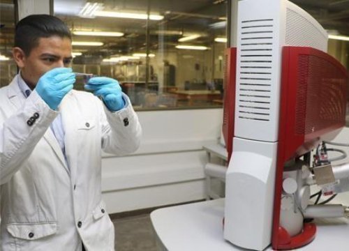 Hombre en laboratorio con guantes y tapabocas mirando fijamente un recipiente de muestreo